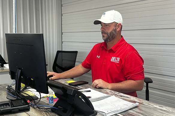 Hunter Myers, Delta Ag team member, working at his desk in a red Delta Ag polo and white cap, focused on his computer in an office environment.
