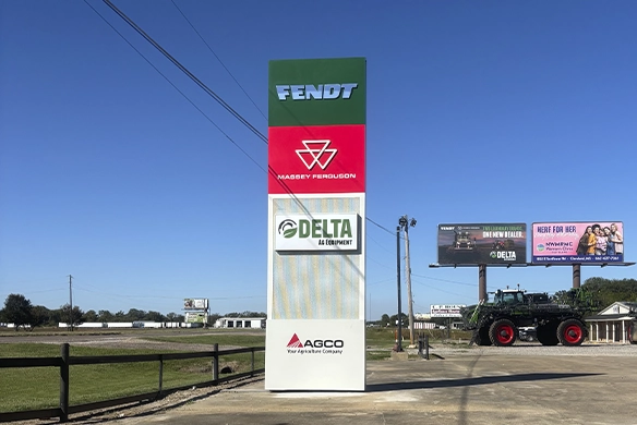 Tall roadside sign displaying Fendt, Massey Ferguson, Delta Ag Equipment, and AGCO logos against a clear blue sky, with a large tractor and billboard in the distance.